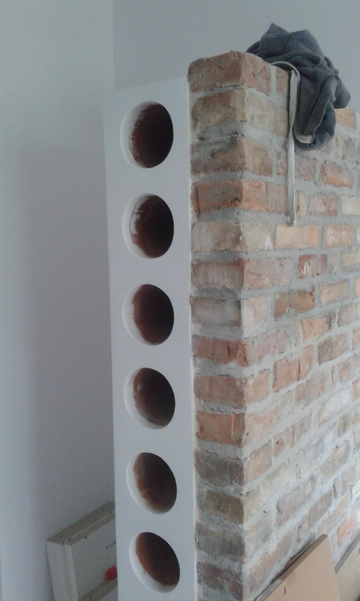 Narożnik pokoju z fragmentem ściany z cegły i białą ścianką działową z okrągłymi otworami, na ceglanej ścianie leży szara bluza z kapturem.