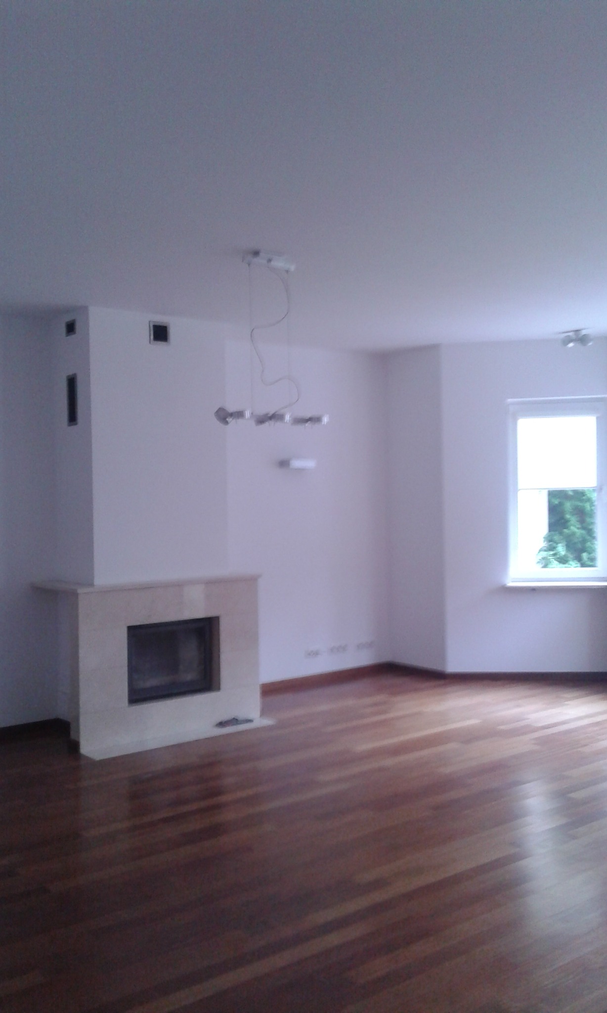 Jasne wnętrze pokoju z parkietem, widocznym kominkiem z beżowego kamienia i nowoczesną lampą wiszącą. Świeżo wykończone, puste pomieszczenie z oknem.