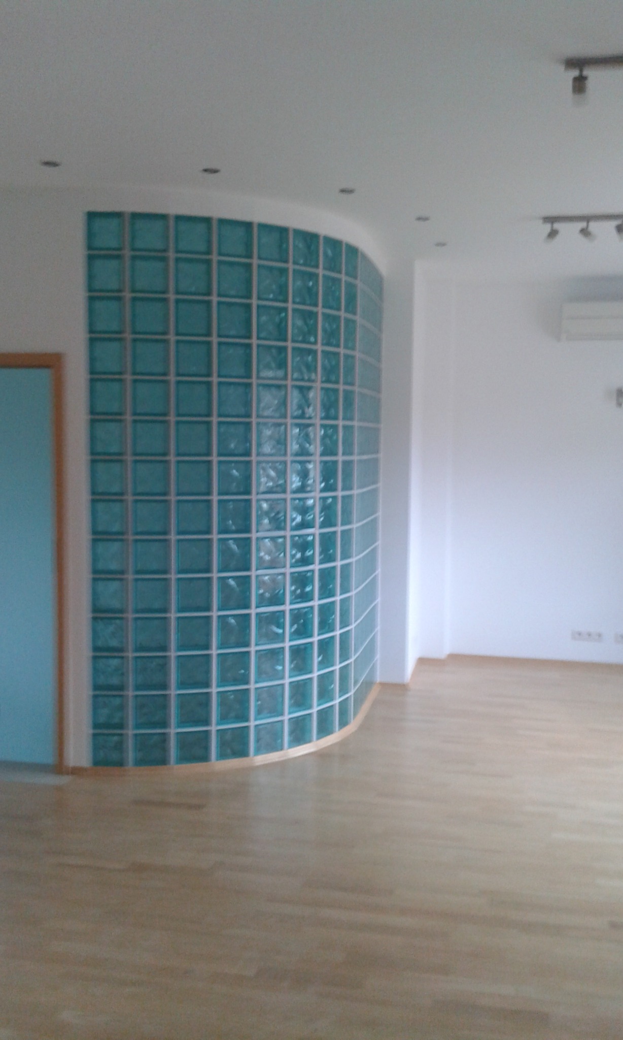 Zakrzywiona ściana z luksferów w odcieniach turkusu, oddzielająca przestrzeń w jasnym, nowoczesnym wnętrzu z parkietem.