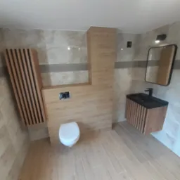 Nowoczesna łazienka z beżowymi płytkami imitującymi drewno, wisząca szafka z umywalką o czarnym blacie, toaleta oraz dekoracyjny grzejnik panelowy z drewnianymi lamelami.