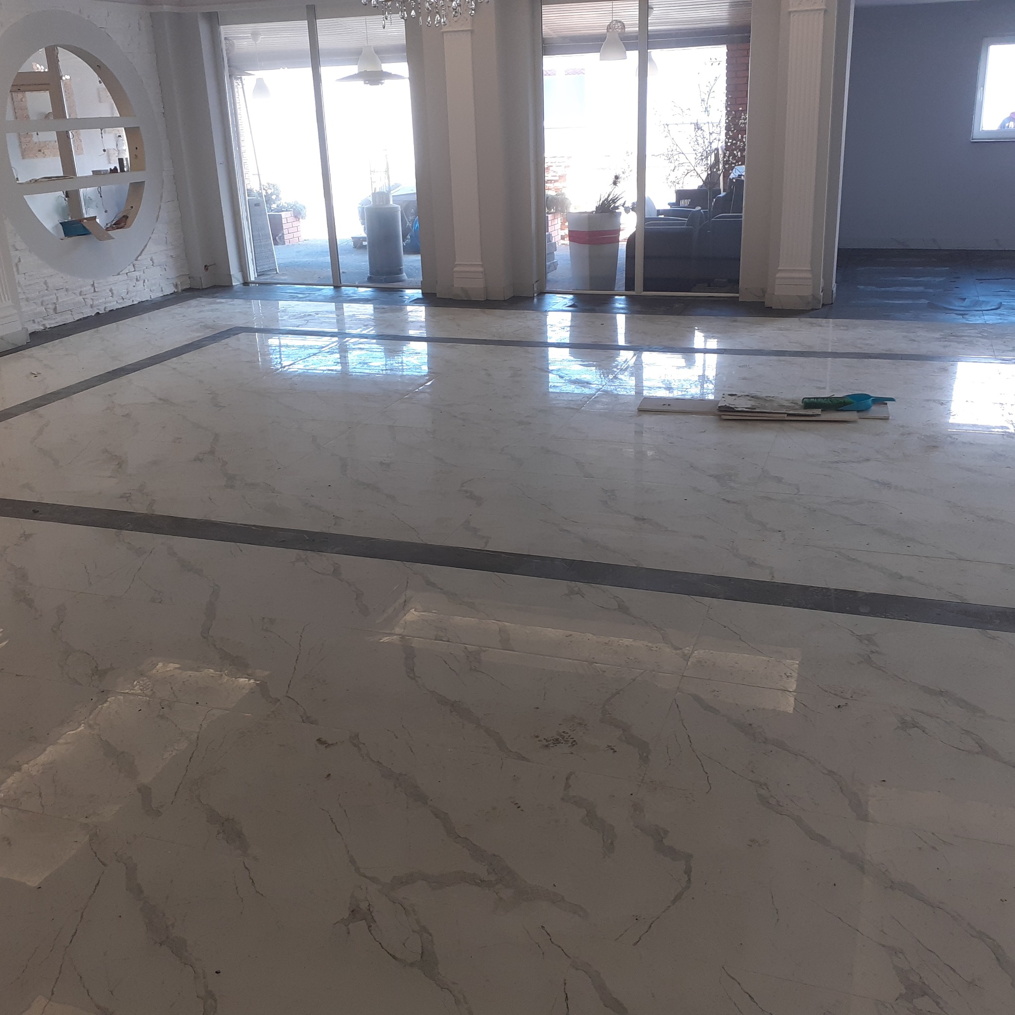 Błyszcząca marmurowa podłoga z ciemnoszarym obramowaniem w przestronnym holu, widoczne narzędzia do czyszczenia podłogi oraz okrągła wnęka w ścianie z półkami.