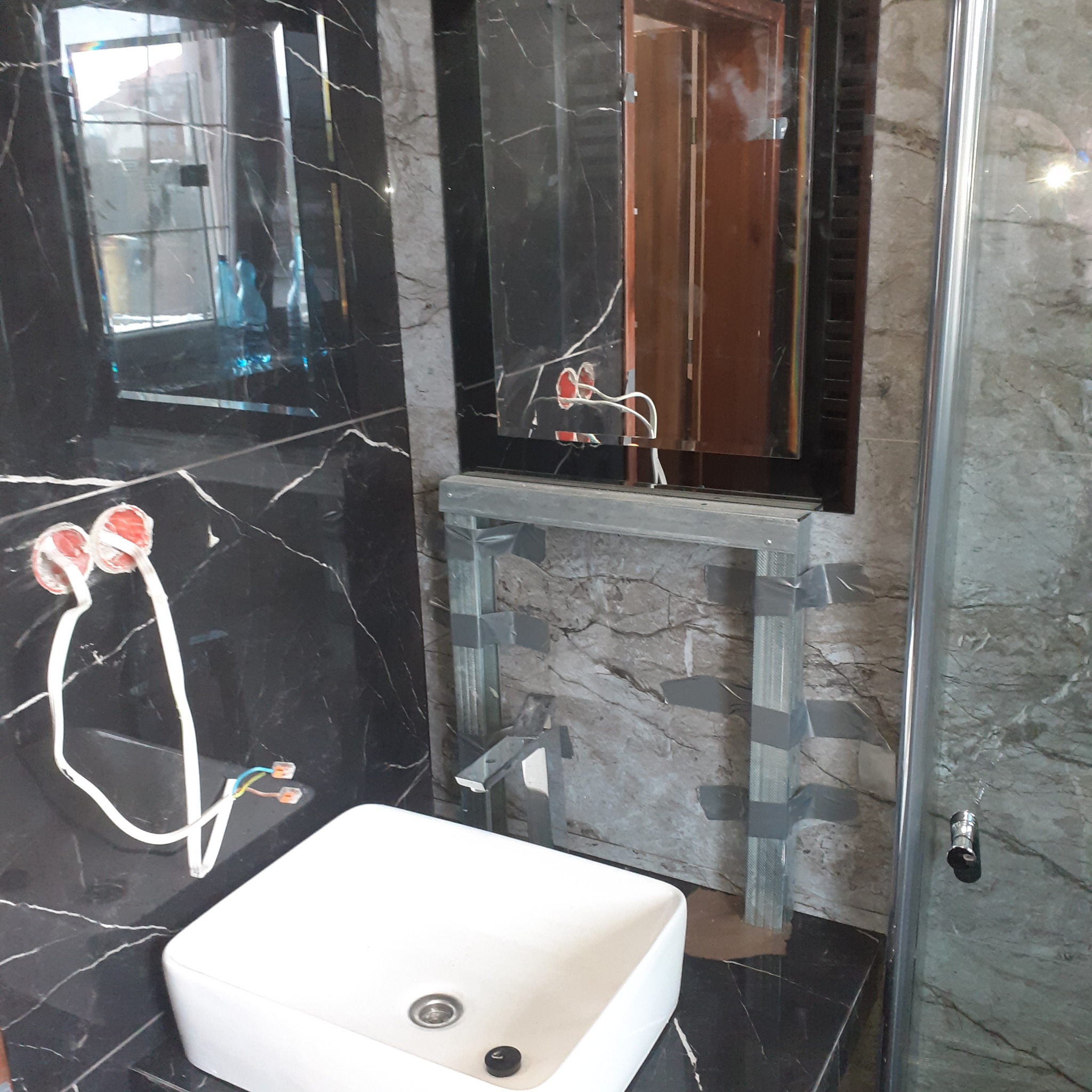 Łazienka w trakcie wykańczania: biała umywalka nablatowa, czarny marmur na ścianach z widocznymi puszkami elektrycznymi, metalowy stelaż pod lustro przymocowany szarą taśmą, kabina prysznicowa w tle.
