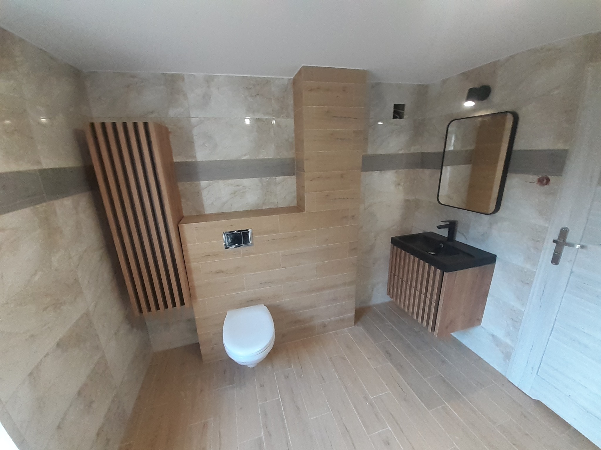 Nowoczesna łazienka z beżowymi płytkami imitującymi drewno, wisząca szafka z umywalką o czarnym blacie, toaleta oraz dekoracyjny grzejnik panelowy z drewnianymi lamelami.