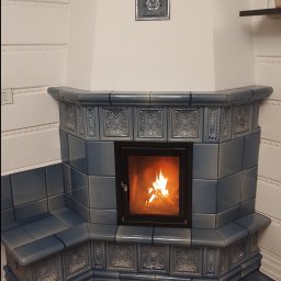 Ceramika babigórska kafle suwaj - Narożny piec kaflowy w odcieniach błękitu z paleniskiem, dekoracyjne kafle z reliefami, drewniana podłoga z efektem postarzania.