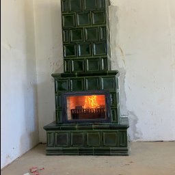 Ceramika babigórska kafle suwaj - Zielony, kaflowy piec kominkowy z palącym się ogniem, w surowym wnętrzu z niedokończonymi ścianami i resztkami materiałów budowlanych na podłodze.