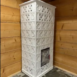 Biały, kaflowy piec z dekoracyjnymi wzorami w rustykalnym wnętrzu z drewnianymi ścianami i kamienną podłogą, zabezpieczony folią ochronną.