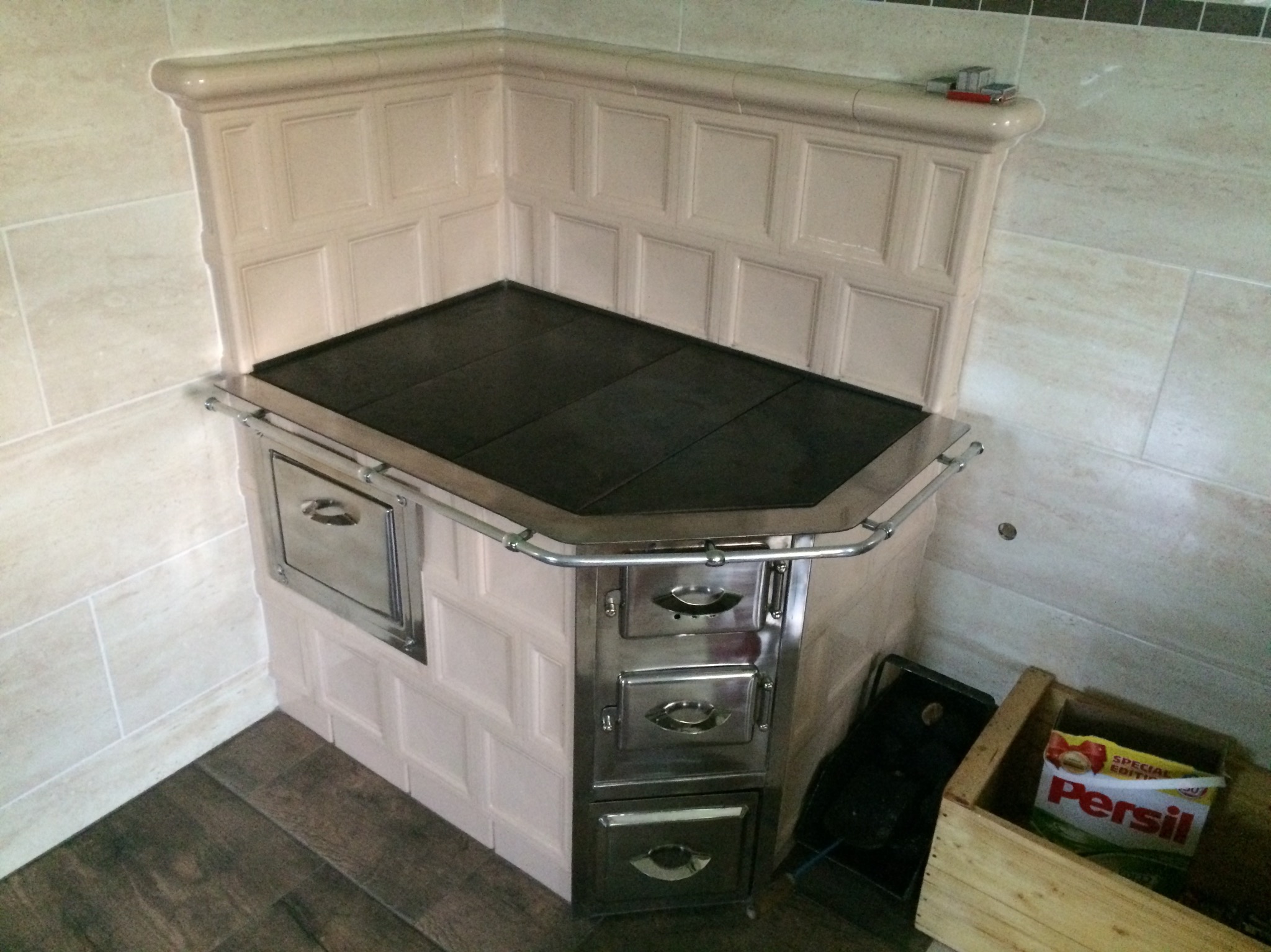 Kuchenka kaflowa z piekarnikiem i płytą grzewczą, wykończona beżowymi kaflami z dekoracyjnymi wgłębieniami, chromowane elementy wykończeniowe, drewniana podłoga i jasne płytki na ścianach.