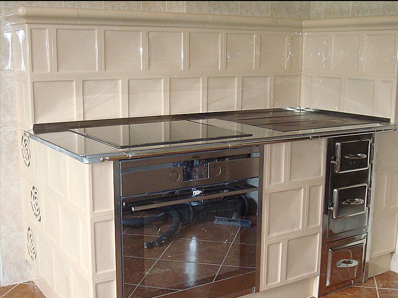 Beżowy piec kaflowy z piekarnikiem i płytą grzewczą, wykończony błyszczącymi kaflami w kwadraty, z widocznym odbiciem otoczenia w szklanych elementach.