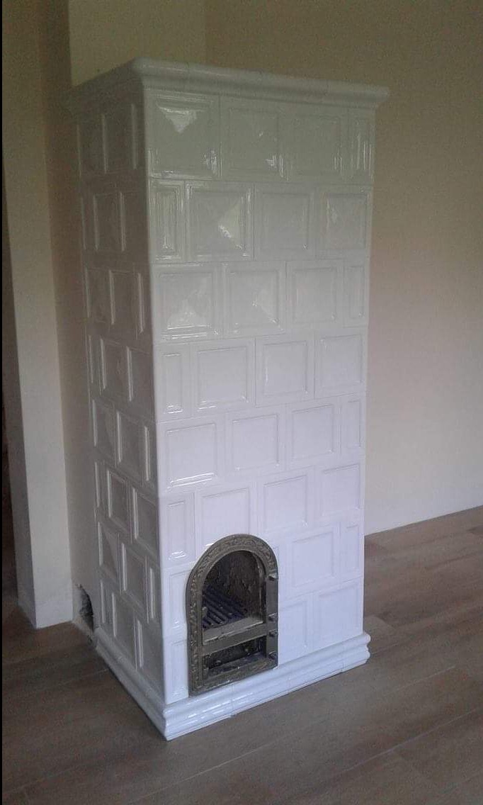 Biały piec kaflowy z metalowymi drzwiczkami paleniska, widoczne niedoskonałości wykończenia przy podłodze.