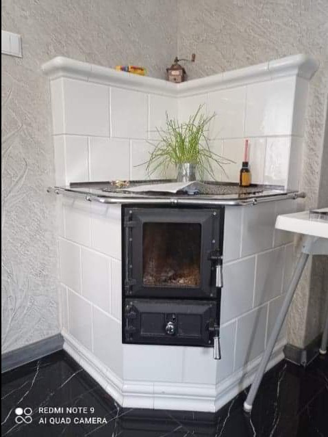 Biały, narożny piec kaflowy z czarnymi drzwiczkami, na blacie roślina w metalowym wiaderku, obok butelka z ciemnym płynem, w tle ściana o fakturze baranka.