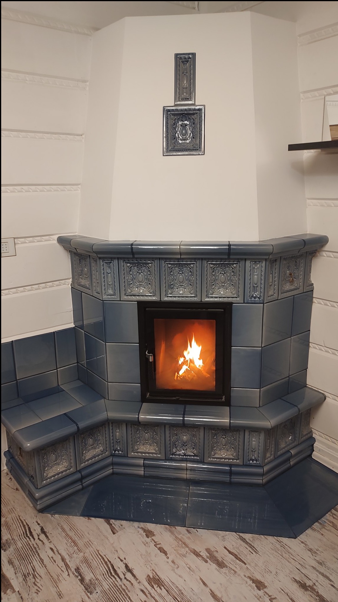 Narożny piec kaflowy w odcieniach błękitu z paleniskiem, dekoracyjne kafle z reliefami, drewniana podłoga z efektem postarzania.