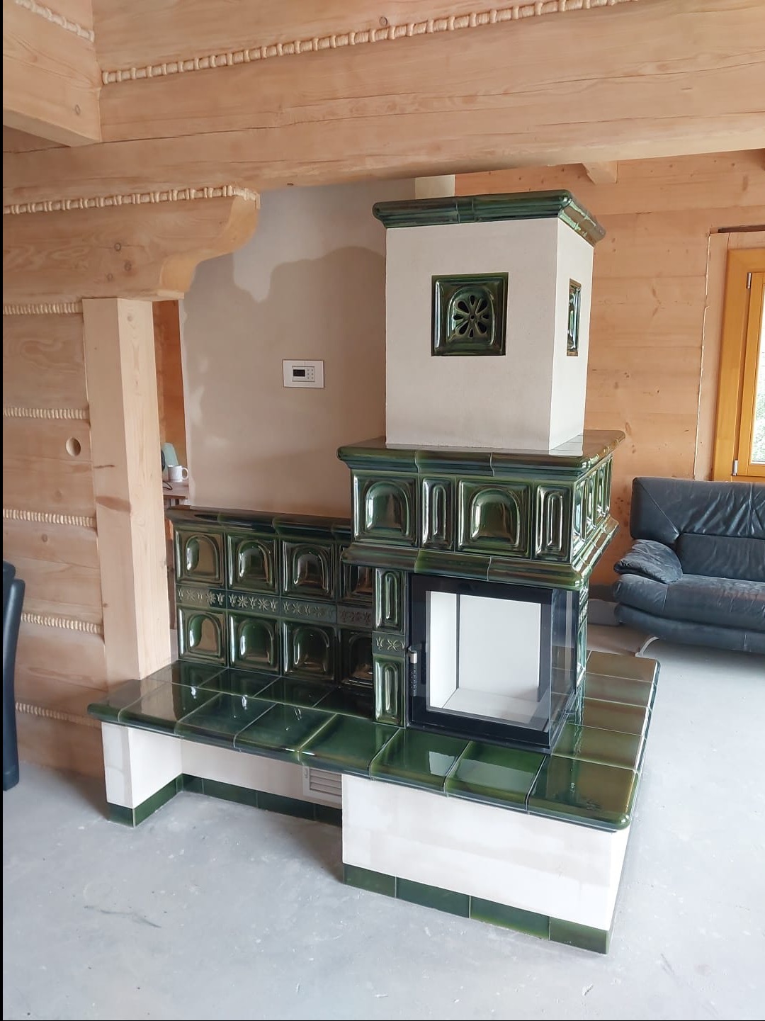 Nowoczesny piec kaflowy z zielonymi kaflami i przeszklonym paleniskiem, w rustykalnym wnętrzu z drewnianymi belkami stropowymi.