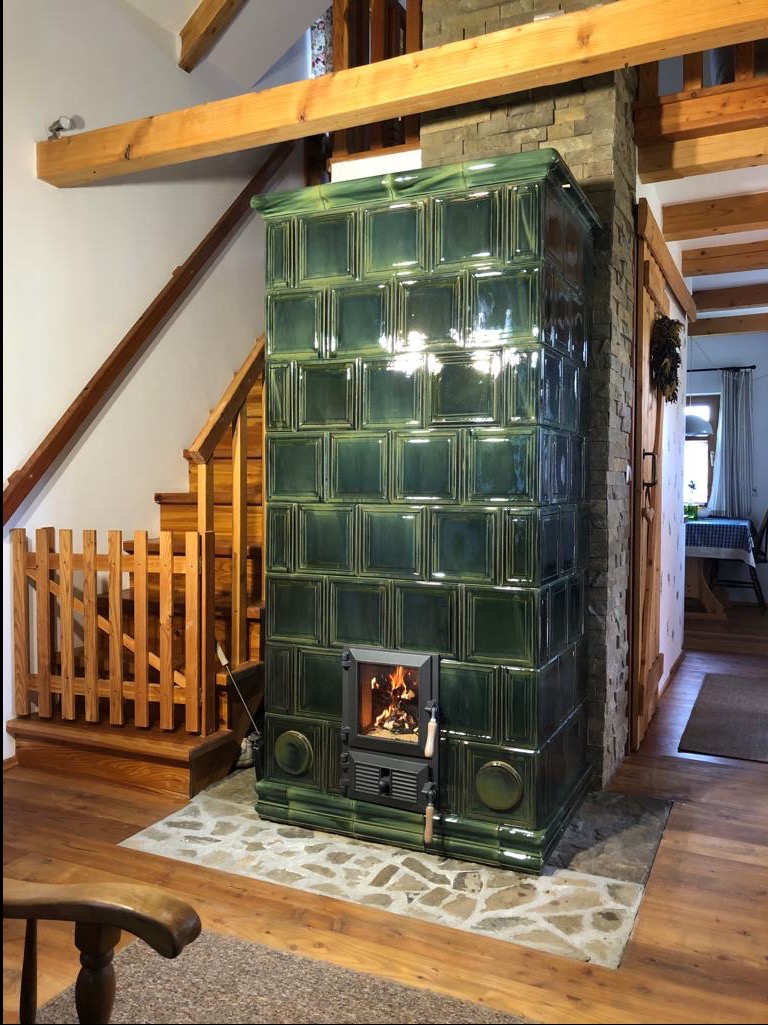 Zielony, kaflowy piec zdobiony kwadratowymi płytkami, z palącym się ogniem w palenisku, w domu w Białce z drewnianymi schodami i elementami wykończenia.