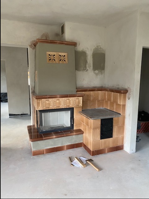 Nowo wybudowany, wykończony brązowymi kaflami piec z żeliwnymi drzwiczkami i blatem w surowym wnętrzu, z widocznymi narzędziami i kawałkami drewna na podłodze.