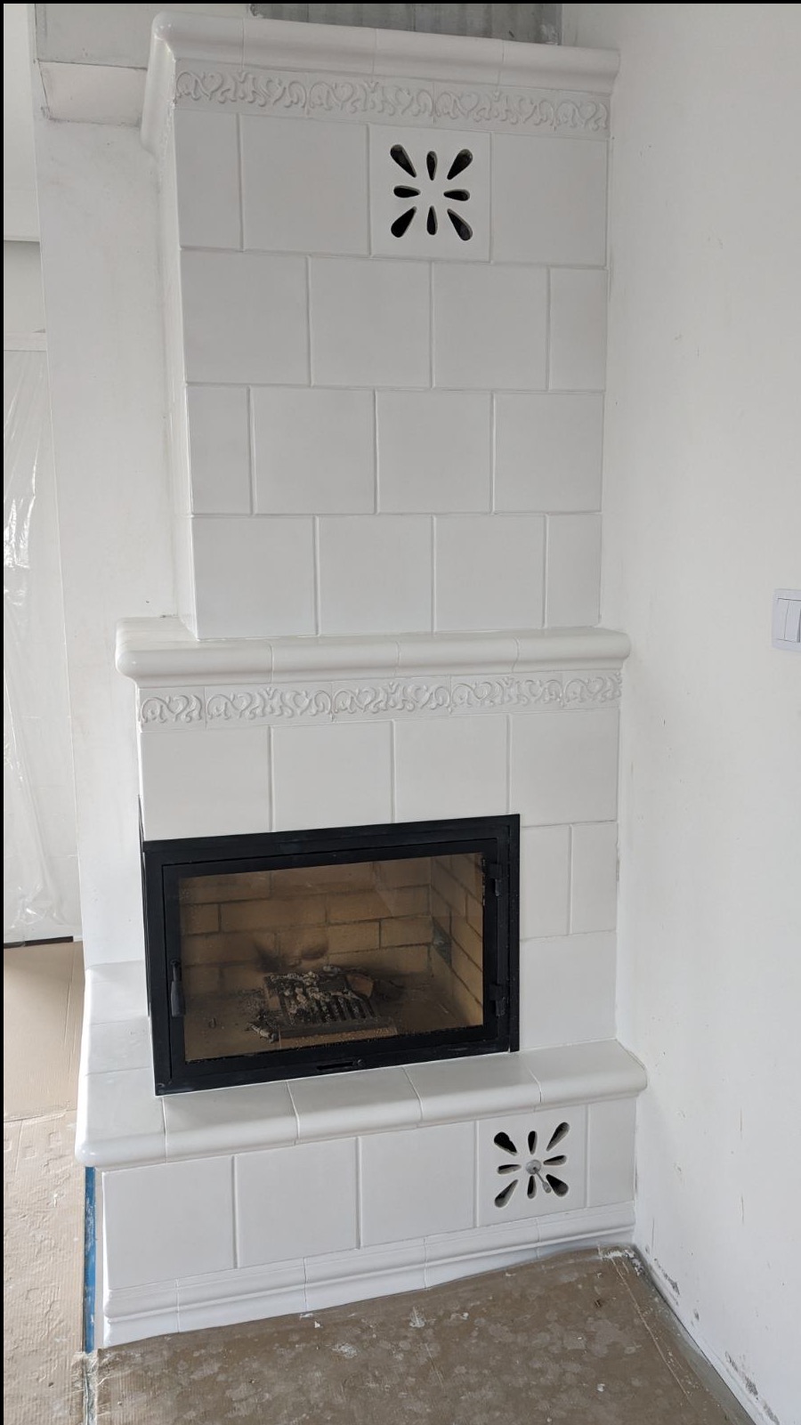 Biały, kafelkowy piec z czarnymi drzwiczkami paleniska i ozdobnymi otworami wentylacyjnymi, widoczny w surowym wnętrzu w trakcie wykańczania.