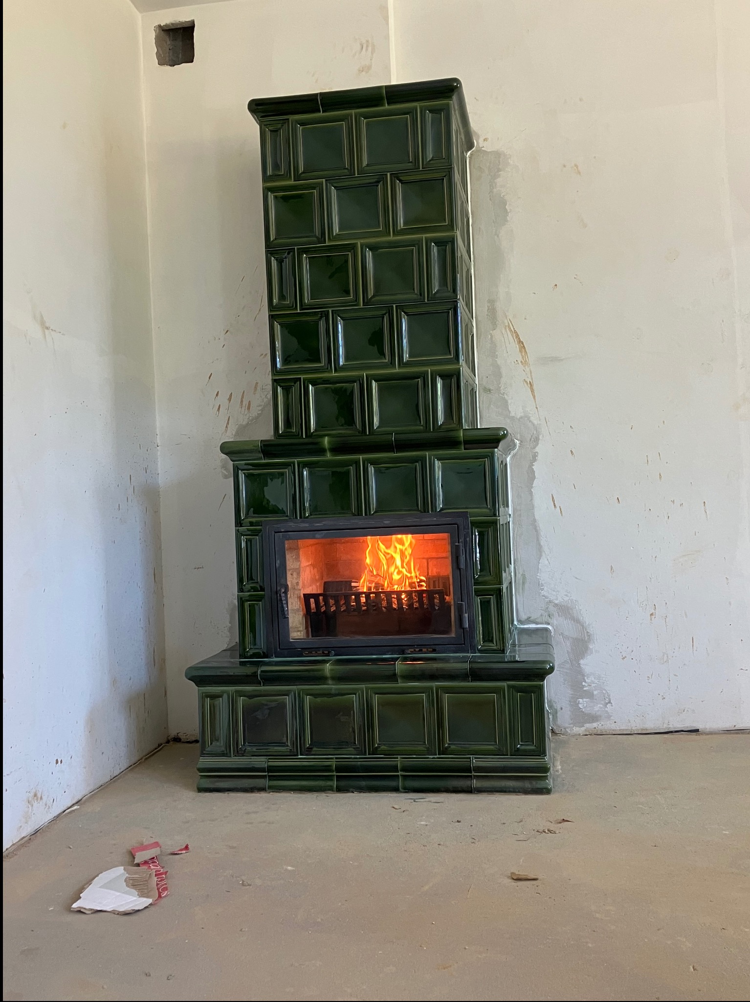 Zielony, kaflowy piec kominkowy z palącym się ogniem, w surowym wnętrzu z niedokończonymi ścianami i resztkami materiałów budowlanych na podłodze.