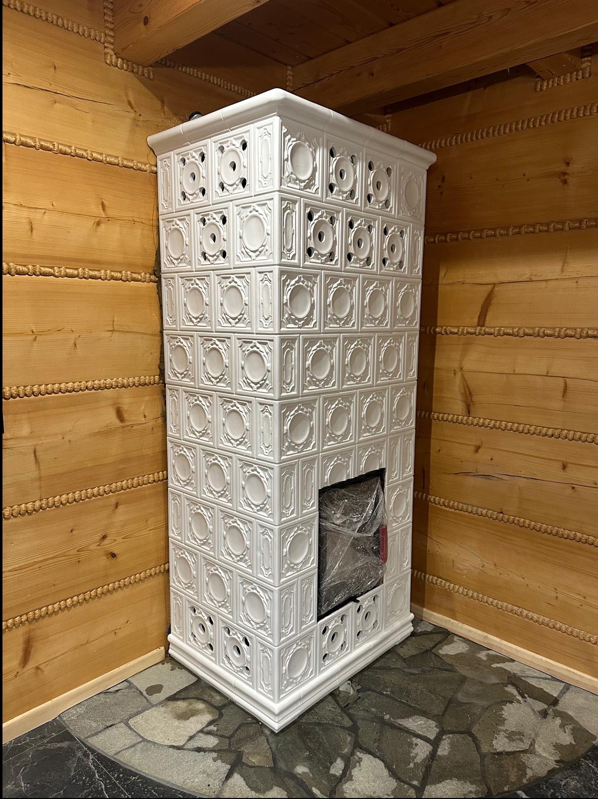 Biały, kaflowy piec z dekoracyjnymi wzorami w rustykalnym wnętrzu z drewnianymi ścianami i kamienną podłogą, zabezpieczony folią ochronną.