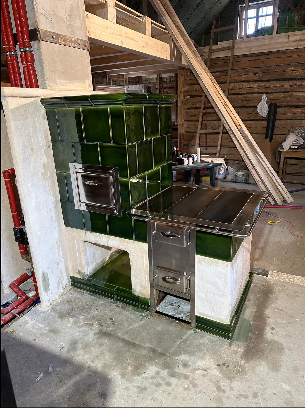 Nowoczesny piec kaflowy z zielonymi kaflami i chromowanymi elementami, w trakcie montażu w surowym wnętrzu drewnianego domu, widoczne instalacje hydrauliczne i rusztowanie.