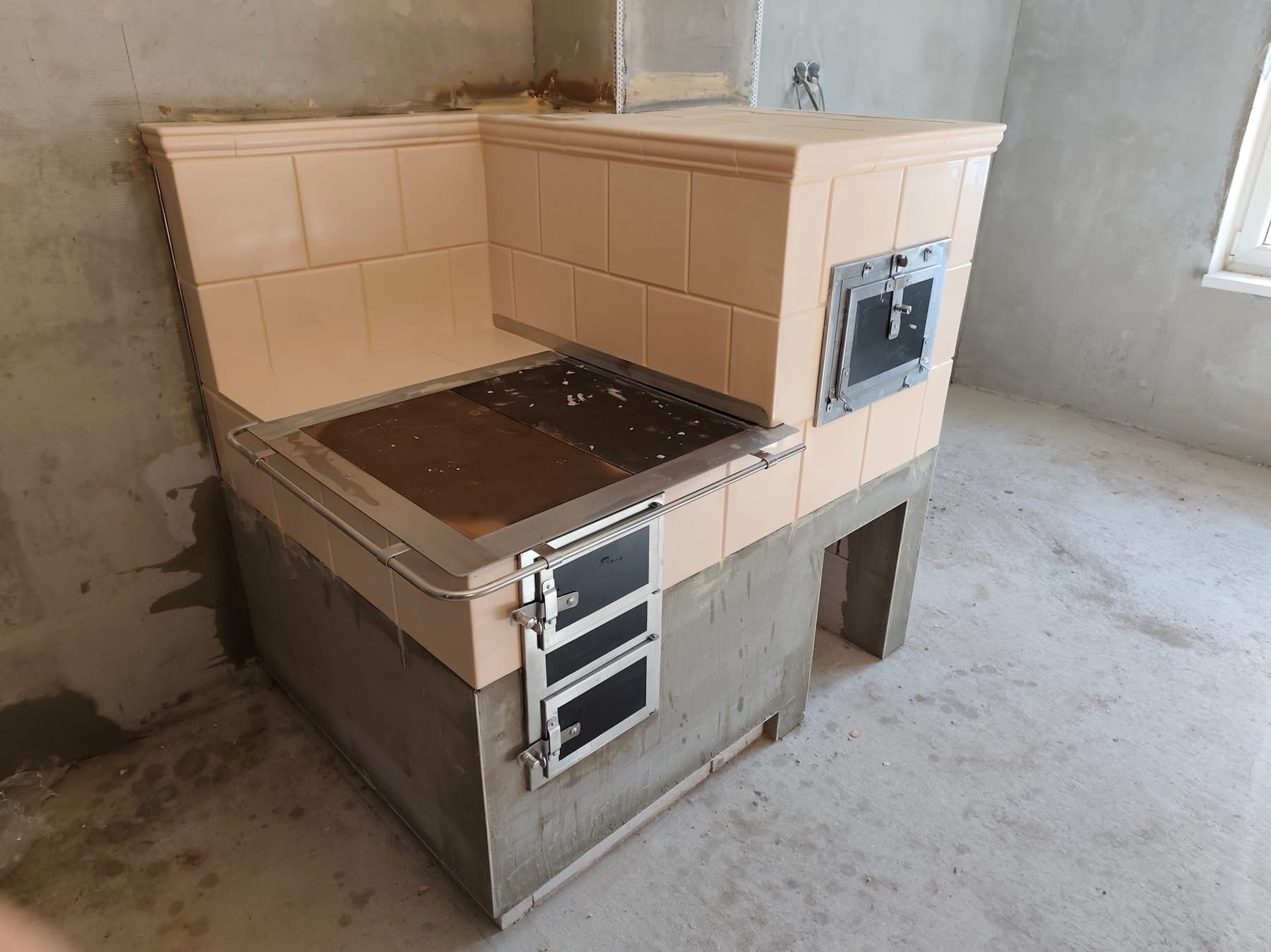 Kuchenna, kaflowa konstrukcja z metalowymi drzwiczkami i blatem, w surowym wnętrzu. Widoczne niedokończone ściany i podłoga. Ujęcie z perspektywy lekko z góry.