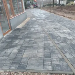 Ułożona szara kostka brukowa prowadząca od domu do drogi, widoczny traktor i wykopana ziemia obok ścieżki.