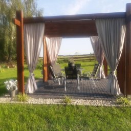 Magiczne Ogrody - Drewniana altana z zasłonami i meblami ogrodowymi na tle zielonego trawnika i drzew