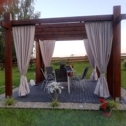 Magiczne Ogrody - Drewniana altana z zasłonami na kostce brukowej otoczonej kamieniami i zielonym trawnikiem, z widocznym zestawem mebli ogrodowych i roślinami w donicach.