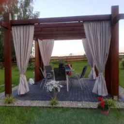 Drewniana altana z zasłonami na kostce brukowej otoczonej kamieniami i zielonym trawnikiem, z widocznym zestawem mebli ogrodowych i roślinami w donicach.