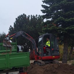 Magiczne Ogrody - Operator w minikoparce załadowuje ziemię do zielonej ciężarówki na tle drzew iglastych i pochmurnego nieba.