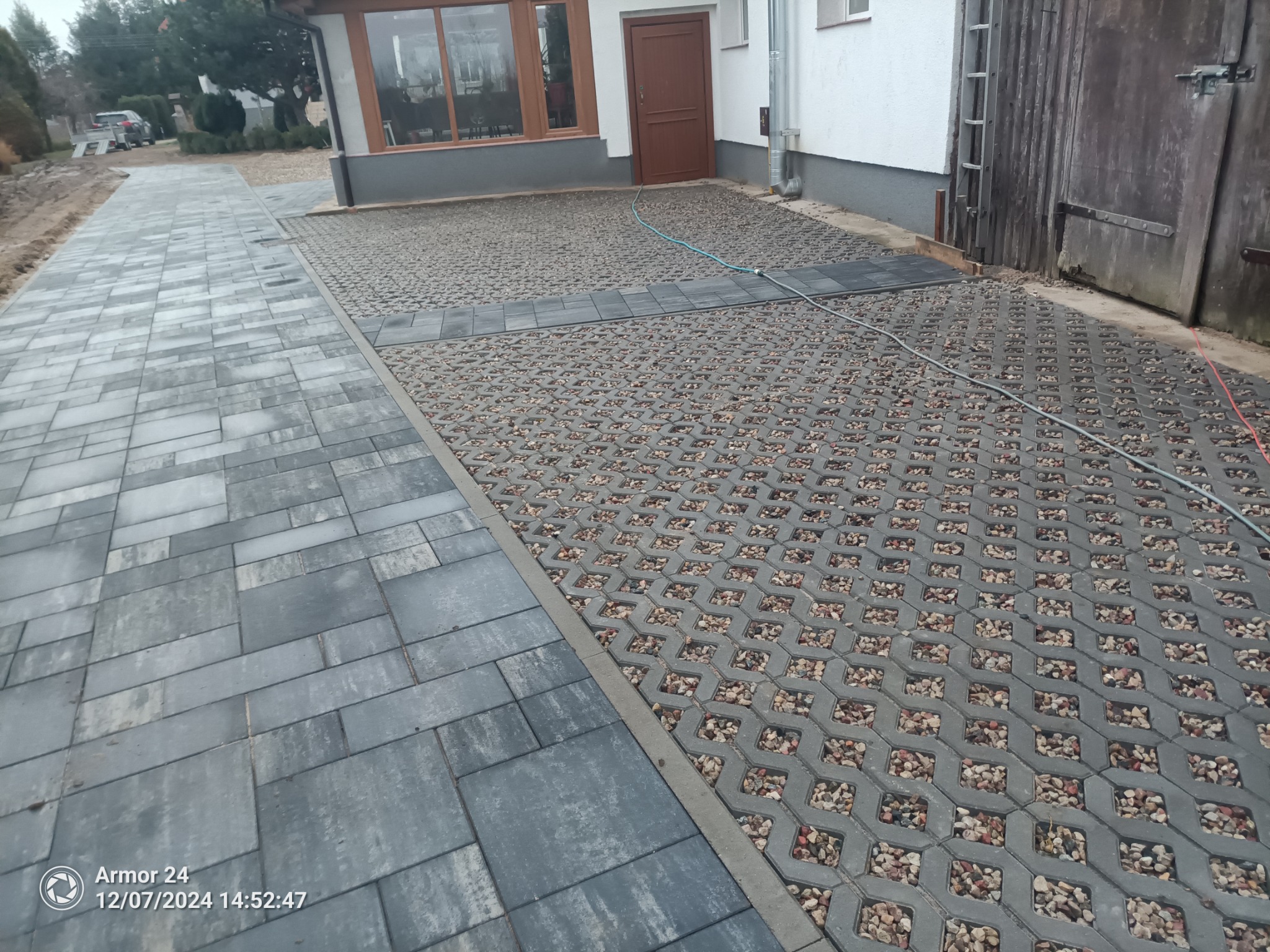 Ułożona nawierzchnia z kostki brukowej i ażurowych płyt wypełnionych drobnym kamieniem, tworząca ścieżkę i podjazd przed budynkiem z brązowymi drzwiami i oknami.