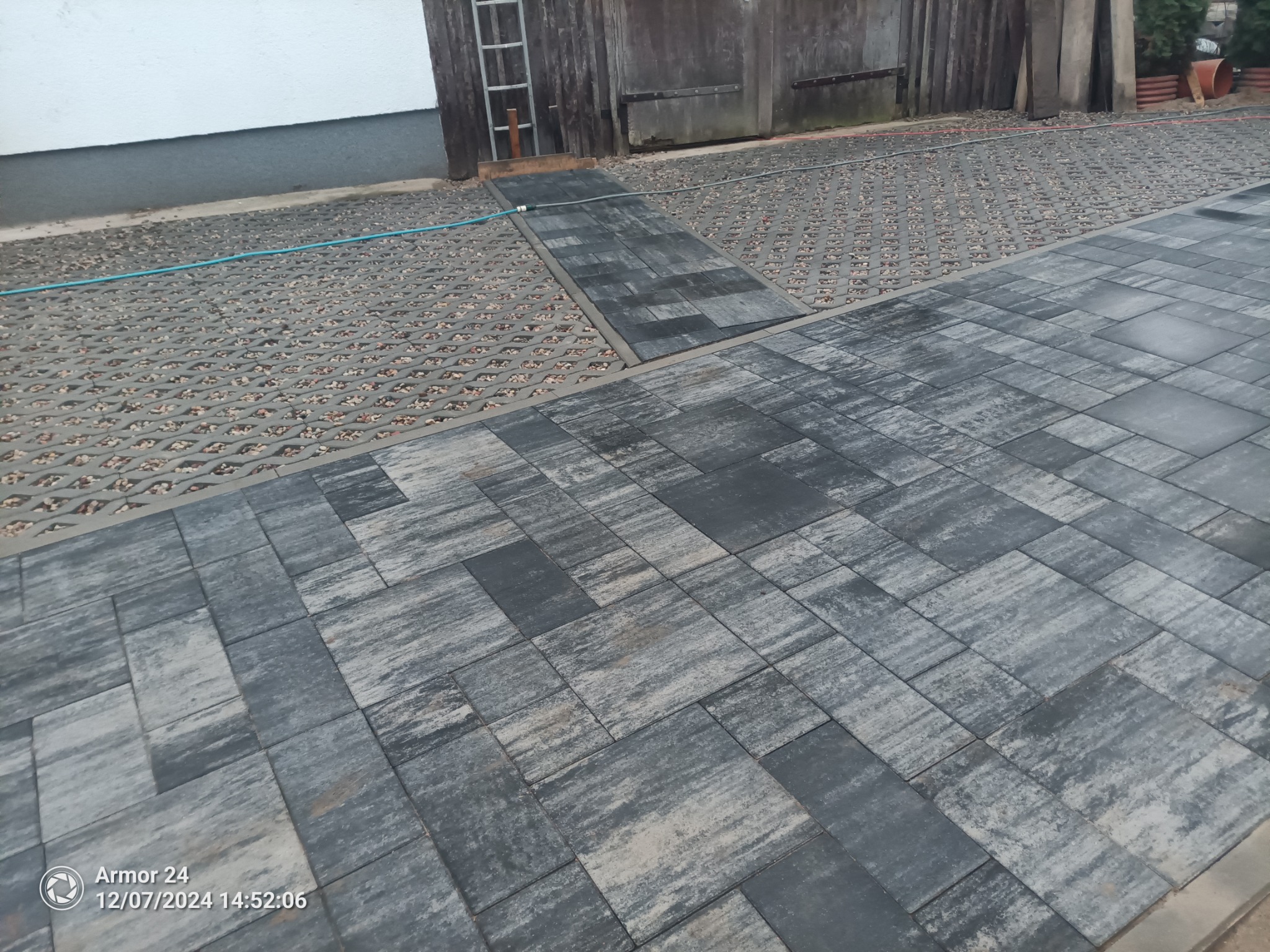 Ułożona szara kostka brukowa tworząca ścieżkę i podjazd z elementami ażurowymi wypełnionymi kamieniami. W tle widoczne drzwi garażowe i fragment ściany budynku.