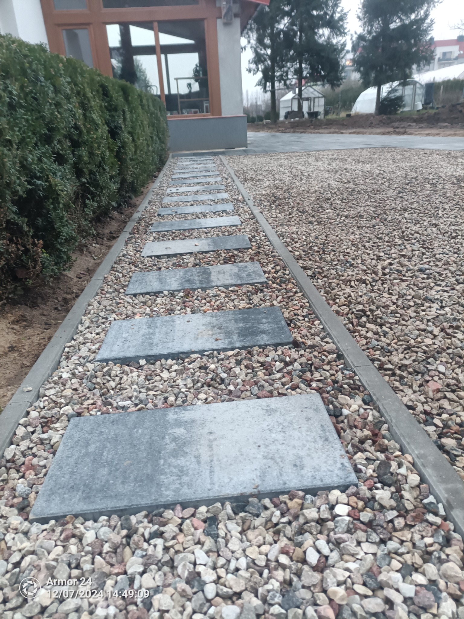 Ścieżka z szarych płyt ułożonych w żwirze z obrzeżami, prowadząca wzdłuż żywopłotu w kierunku domu.