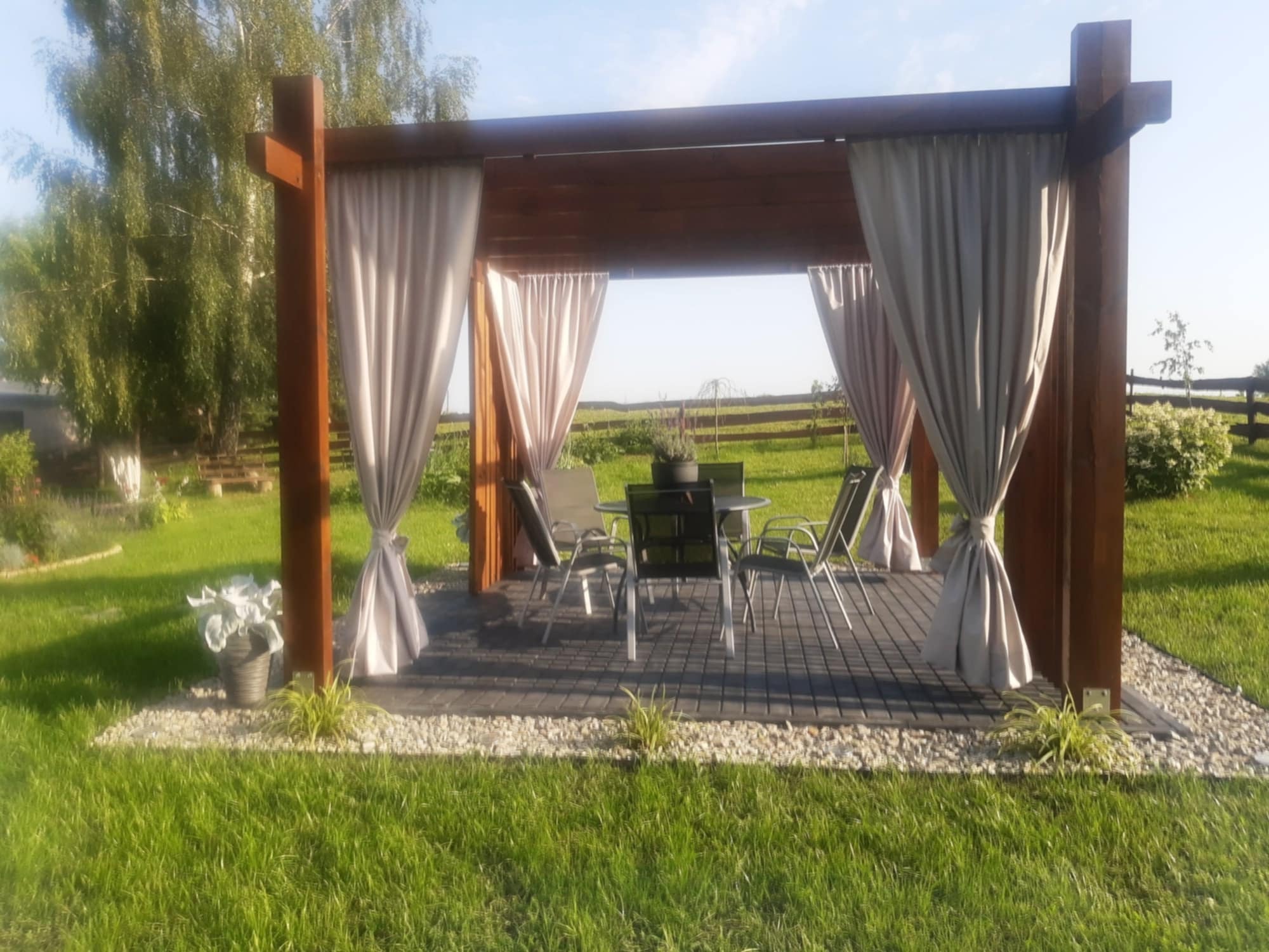 Drewniana altana z zasłonami i meblami ogrodowymi na tle zielonego trawnika i drzew