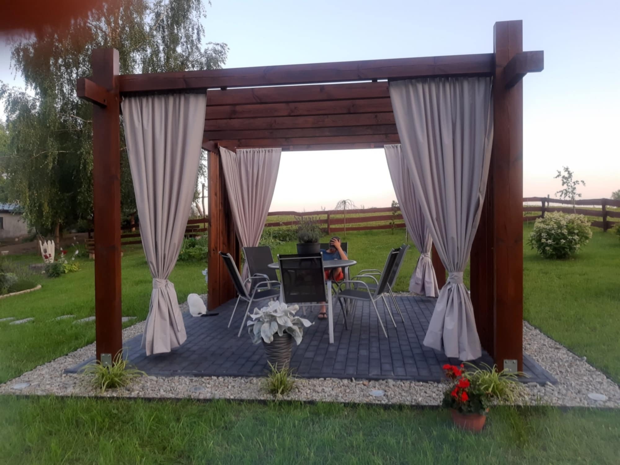Drewniana altana z zasłonami na kostce brukowej otoczonej kamieniami i zielonym trawnikiem, z widocznym zestawem mebli ogrodowych i roślinami w donicach.