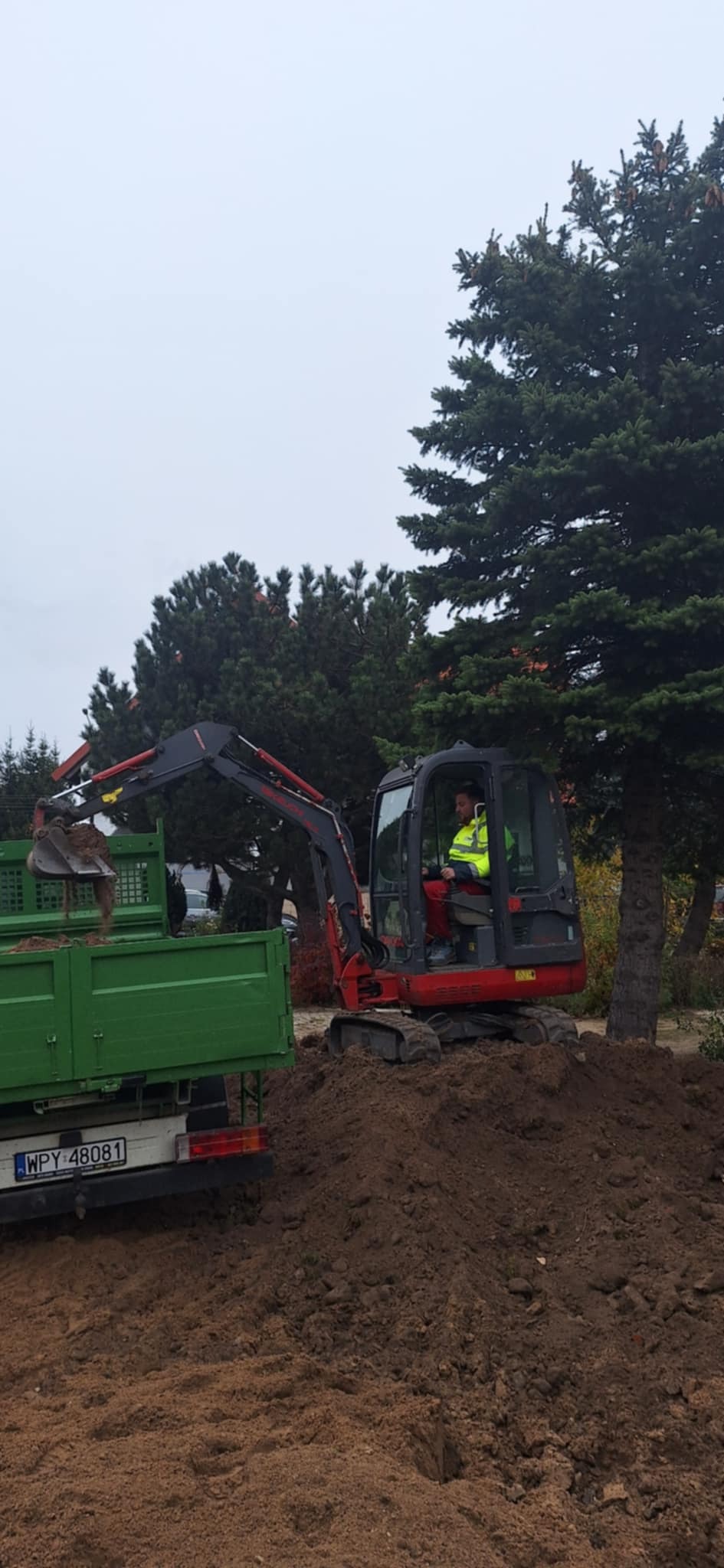 Operator w minikoparce załadowuje ziemię do zielonej ciężarówki na tle drzew iglastych i pochmurnego nieba.