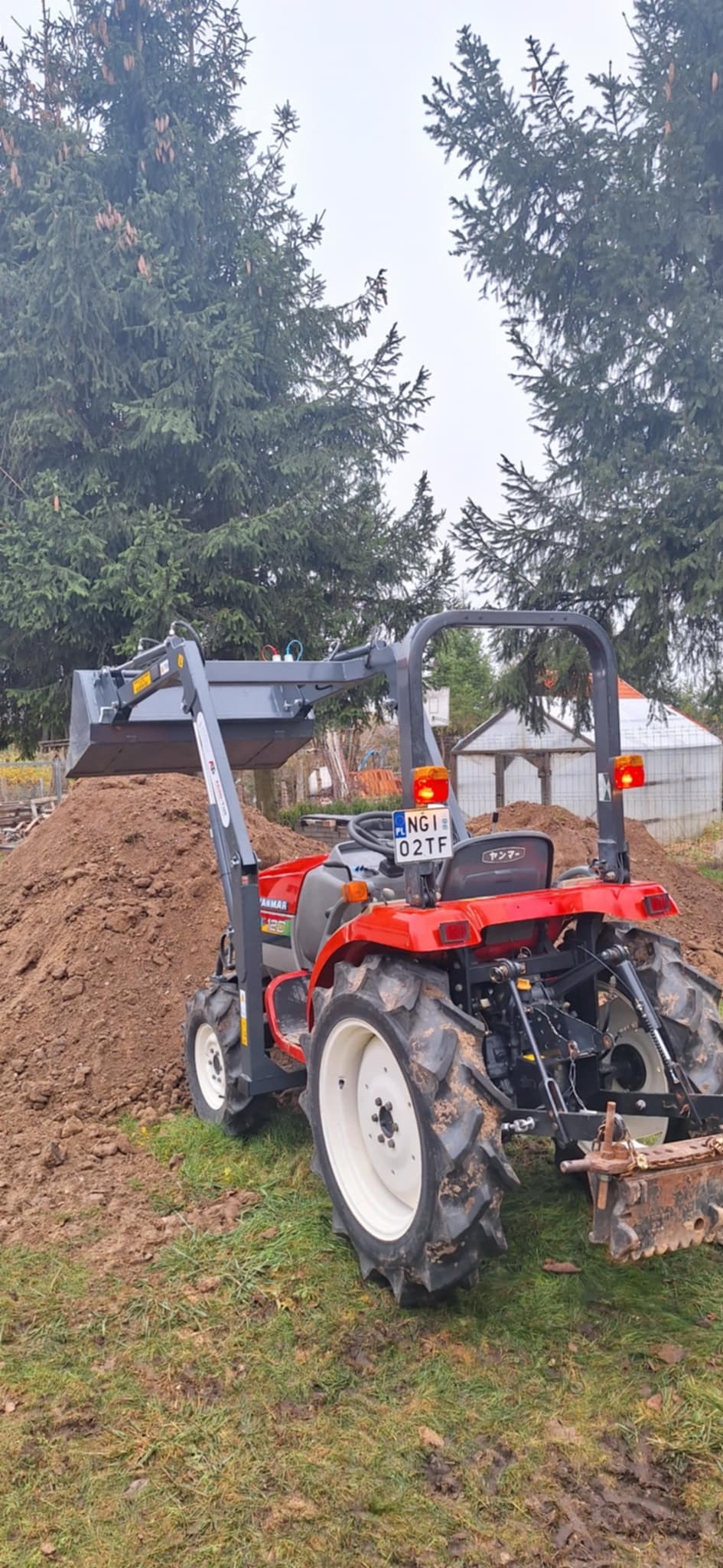 Czerwony traktor Yanmar z szarą łychą na tle sterty ziemi i zielonych drzew iglastych, widok z tyłu, tablica rejestracyjna NGI 02TF.