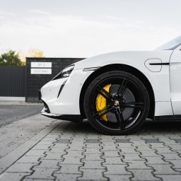 Mąsior Automotive Photographer - Biały Porsche Taycan z czarnymi felgami i żółtymi zaciskami hamulcowymi, sfotografowany z niskiego kąta na tle nowoczesnego ogrodzenia.