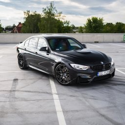 Mąsior Automotive Photographer - Czarny samochód BMW M3 stoi na betonowym parkingu wielopoziomowym z widocznymi śladami opon, w tle drzewa i betonowy mur z oznaczeniami miejsc parkingowych.