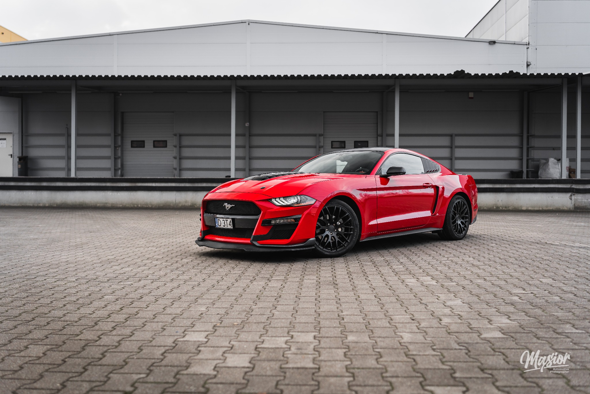 Czerwony Ford Mustang z czarnymi felgami na tle szarego budynku magazynowego, prezentowany z niskiej perspektywy.