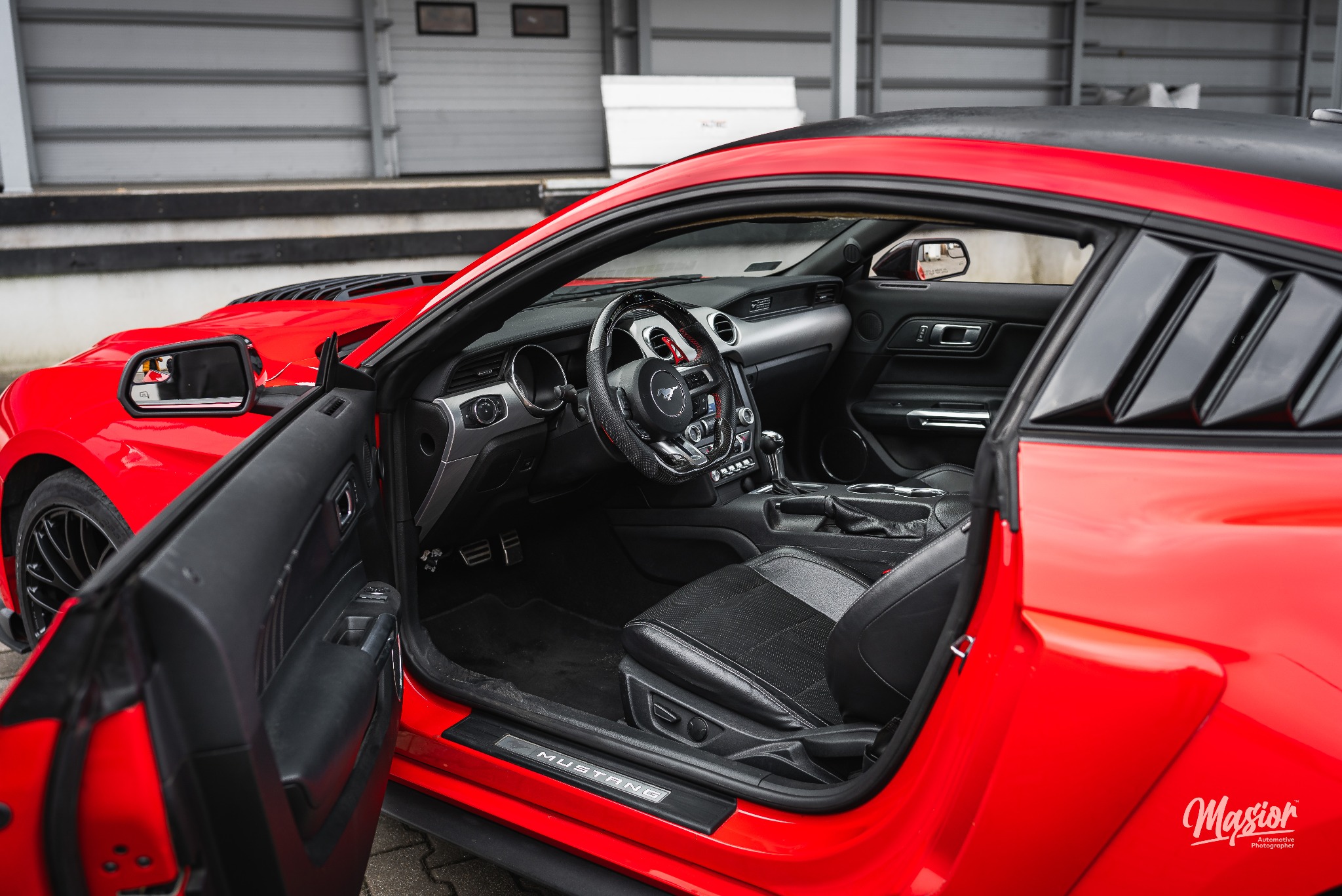 Czerwony Ford Mustang z otwartymi drzwiami, widoczny kokpit z kierownicą obszytą skórą i aluminiowymi detalami, na progu napis 'Mustang', w tle szara ściana garażu.