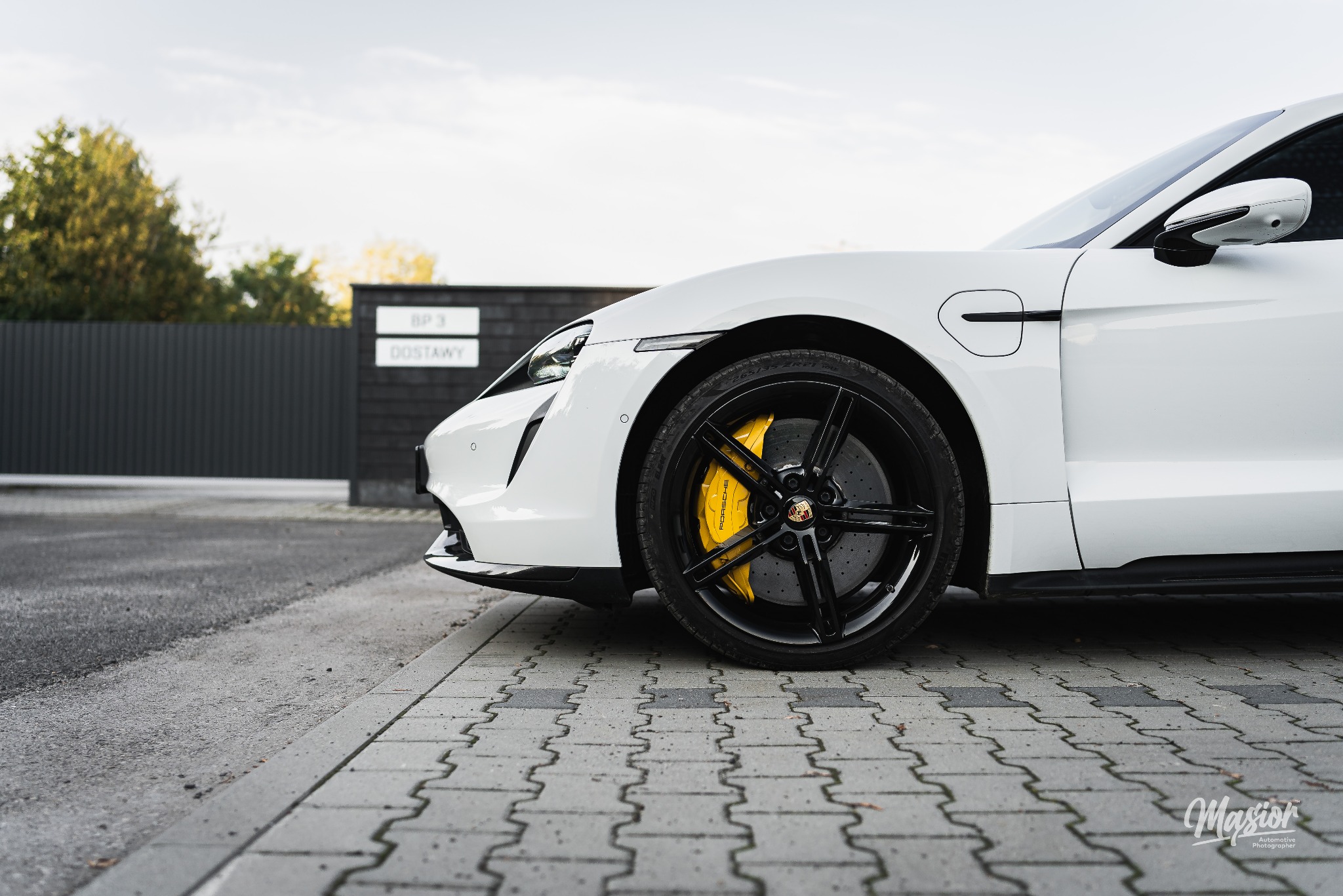 Biały Porsche Taycan z czarnymi felgami i żółtymi zaciskami hamulcowymi, sfotografowany z niskiego kąta na tle nowoczesnego ogrodzenia.