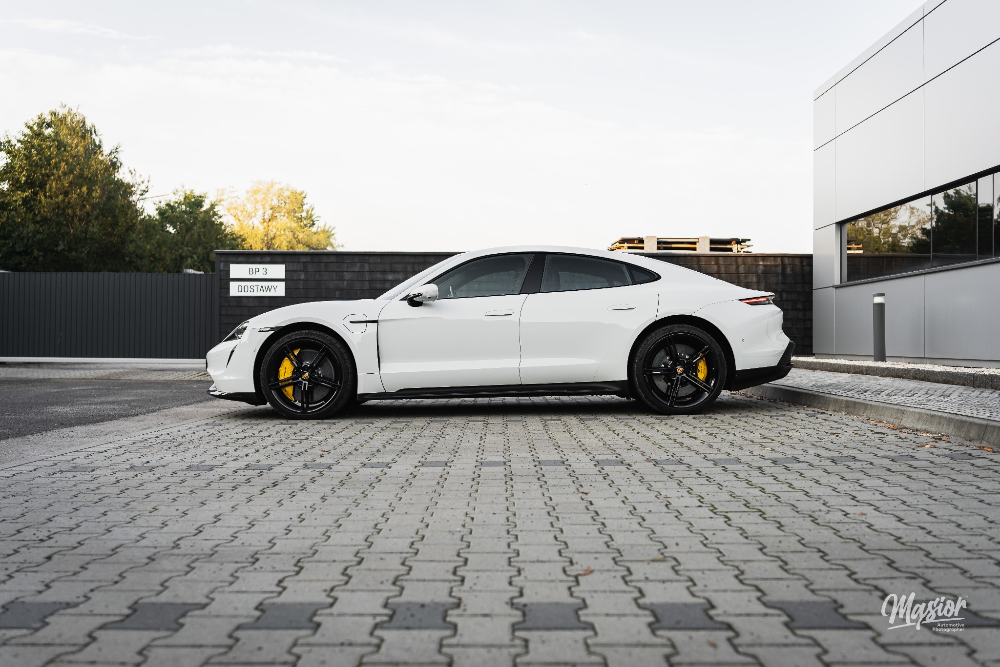 Biały Porsche Taycan z czarnymi felgami i żółtymi zaciskami hamulcowymi, zaparkowany na szarej kostce brukowej przed nowoczesnym budynkiem z szarą ścianą i tabliczką 'BP 3 Dostawy'.