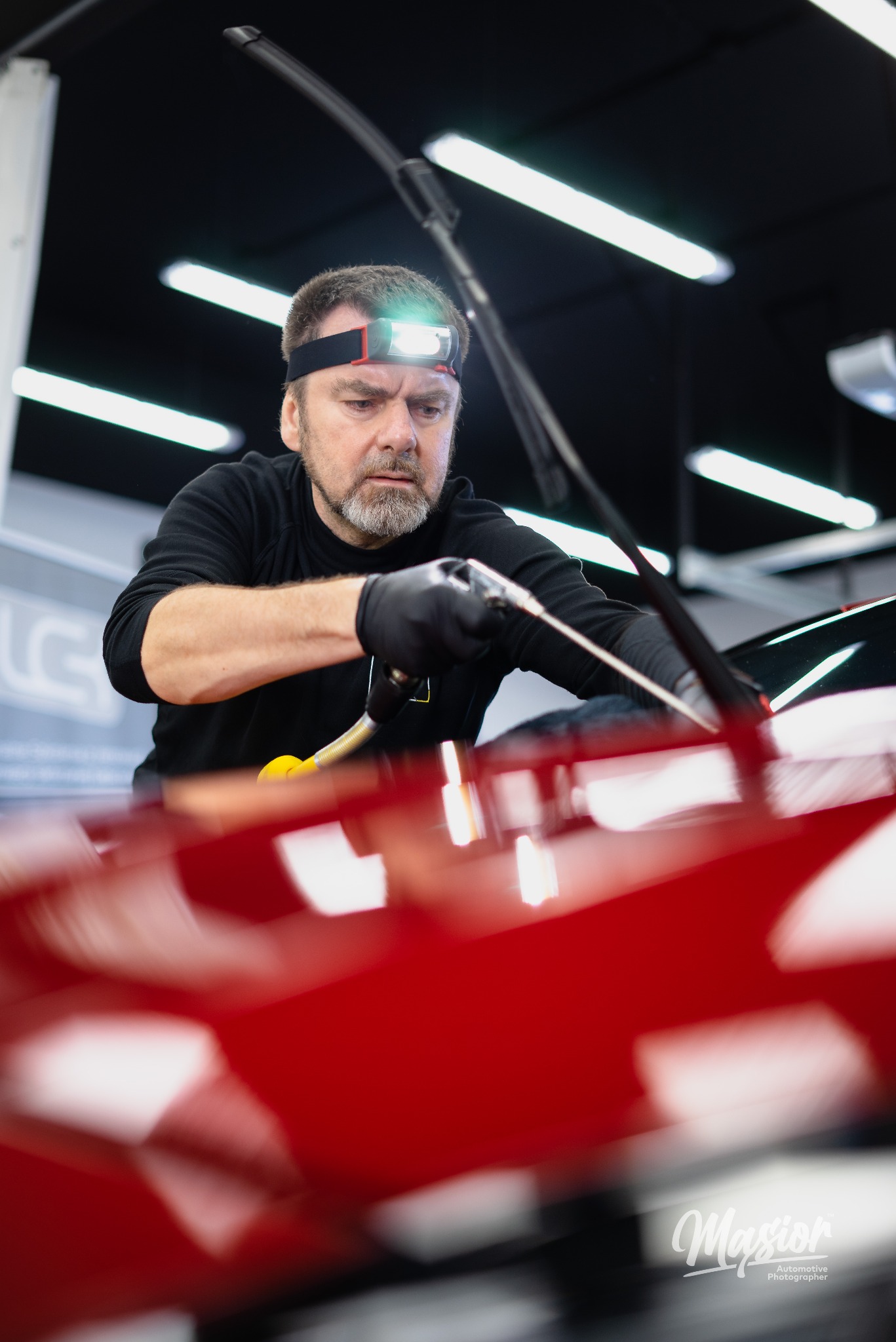 Detailer w czarnych rękawicach i z czołówką, precyzyjnie czyści czerwoną karoserię samochodu za pomocą pistoletu pneumatycznego i mikrofibry, widoczne odbicia lamp w lakierze.