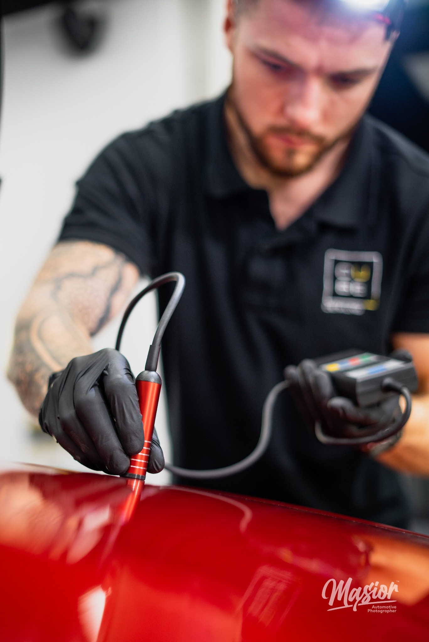 Detailing samochodu: Mężczyzna w czarnych rękawiczkach mierzy grubość lakieru czerwonego auta za pomocą elektronicznego miernika z czerwoną sondą.