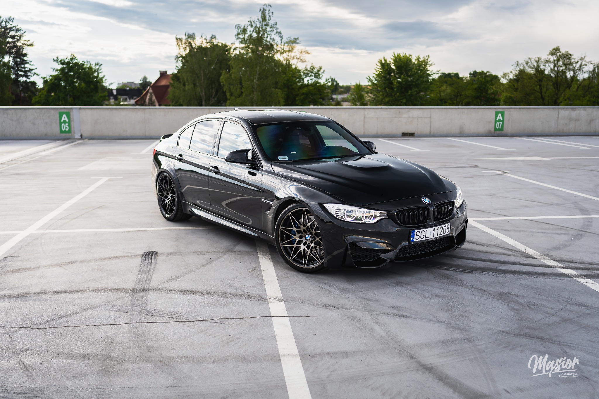Czarny samochód BMW M3 stoi na betonowym parkingu wielopoziomowym z widocznymi śladami opon, w tle drzewa i betonowy mur z oznaczeniami miejsc parkingowych.