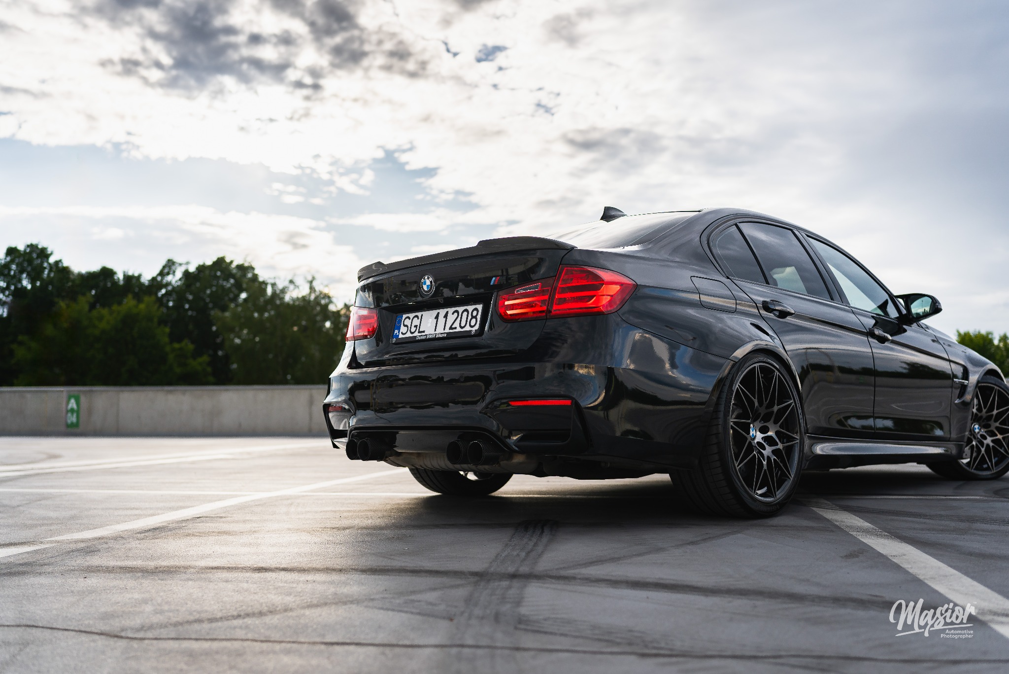 Czarny samochód BMW M3 z karbonowym spojlerem, widok z tyłu na parkingu z śladami opon, tablica rejestracyjna SGL 11208