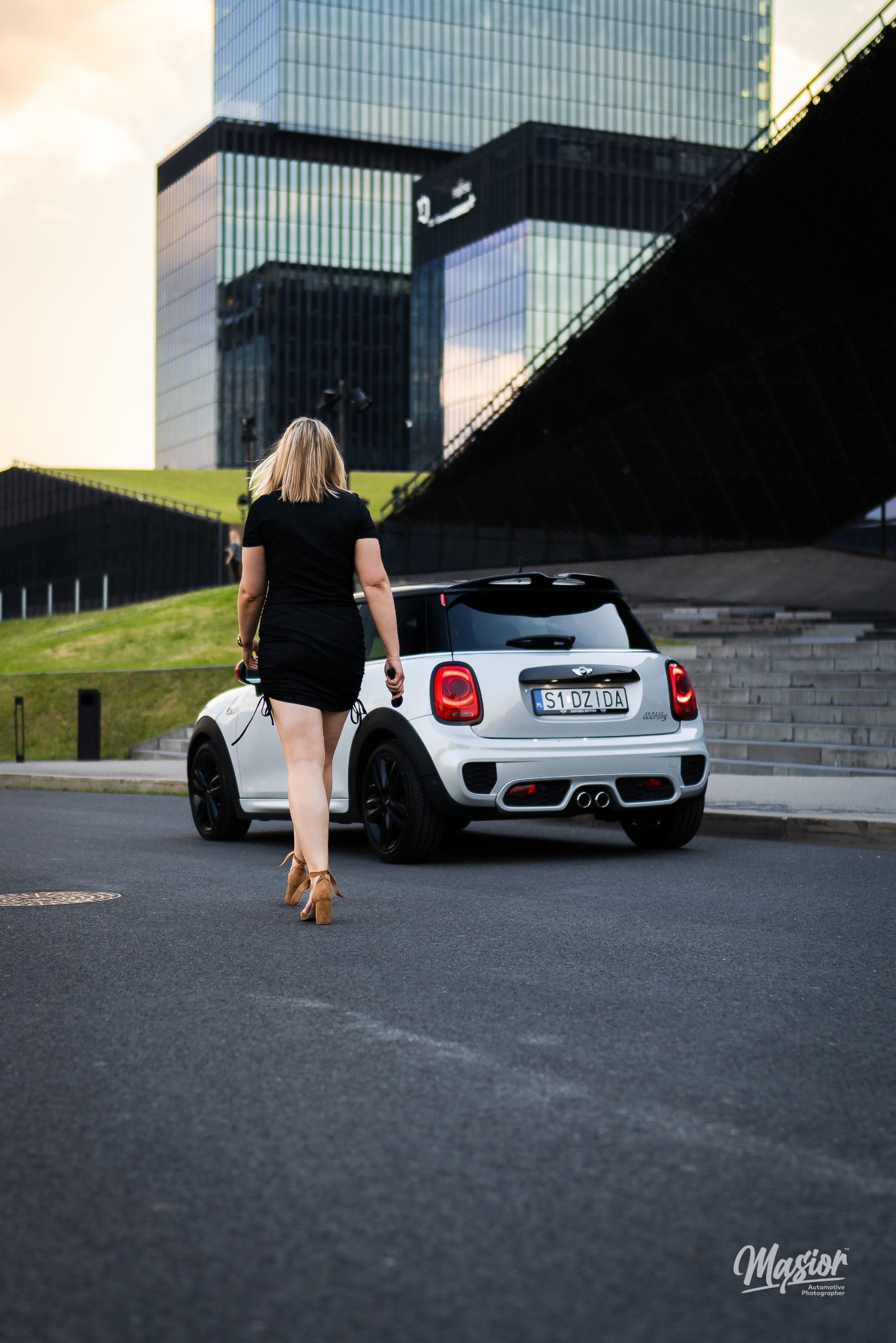 Kobieta w czarnej sukience i brązowych butach na obcasie idzie w kierunku białego Mini Cooper S zaparkowanego na tle nowoczesnej architektury miejskiej.