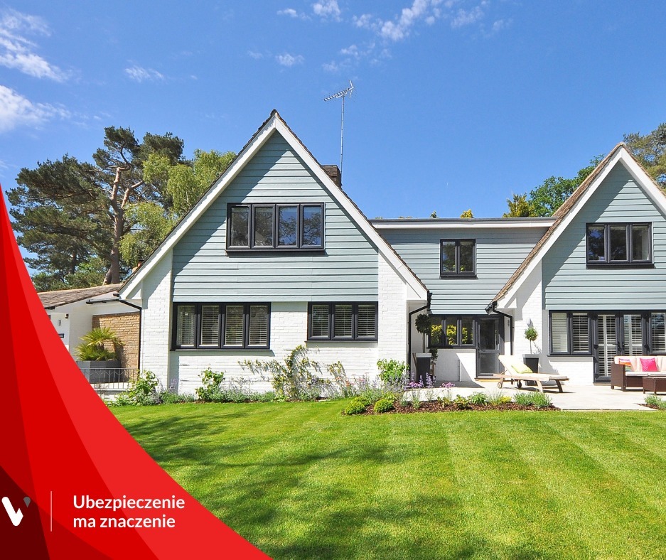 Dwukondygnacyjny dom z szarą elewacją i białymi ścianami, zadbany trawnik, błękitne niebo w tle, antena na dachu, częściowo zasłonięty logo firmy ubezpieczeniowej w lewym dolnym rogu.