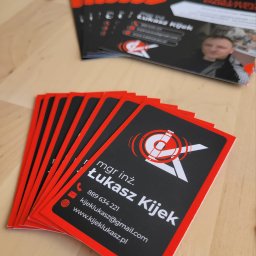 Łukasz Kijek - Stos wizytówek mgr inż. Łukasza Kijka z czarnym tłem, czerwonym obramowaniem i białym logo celownika, leżących na drewnianym blacie.