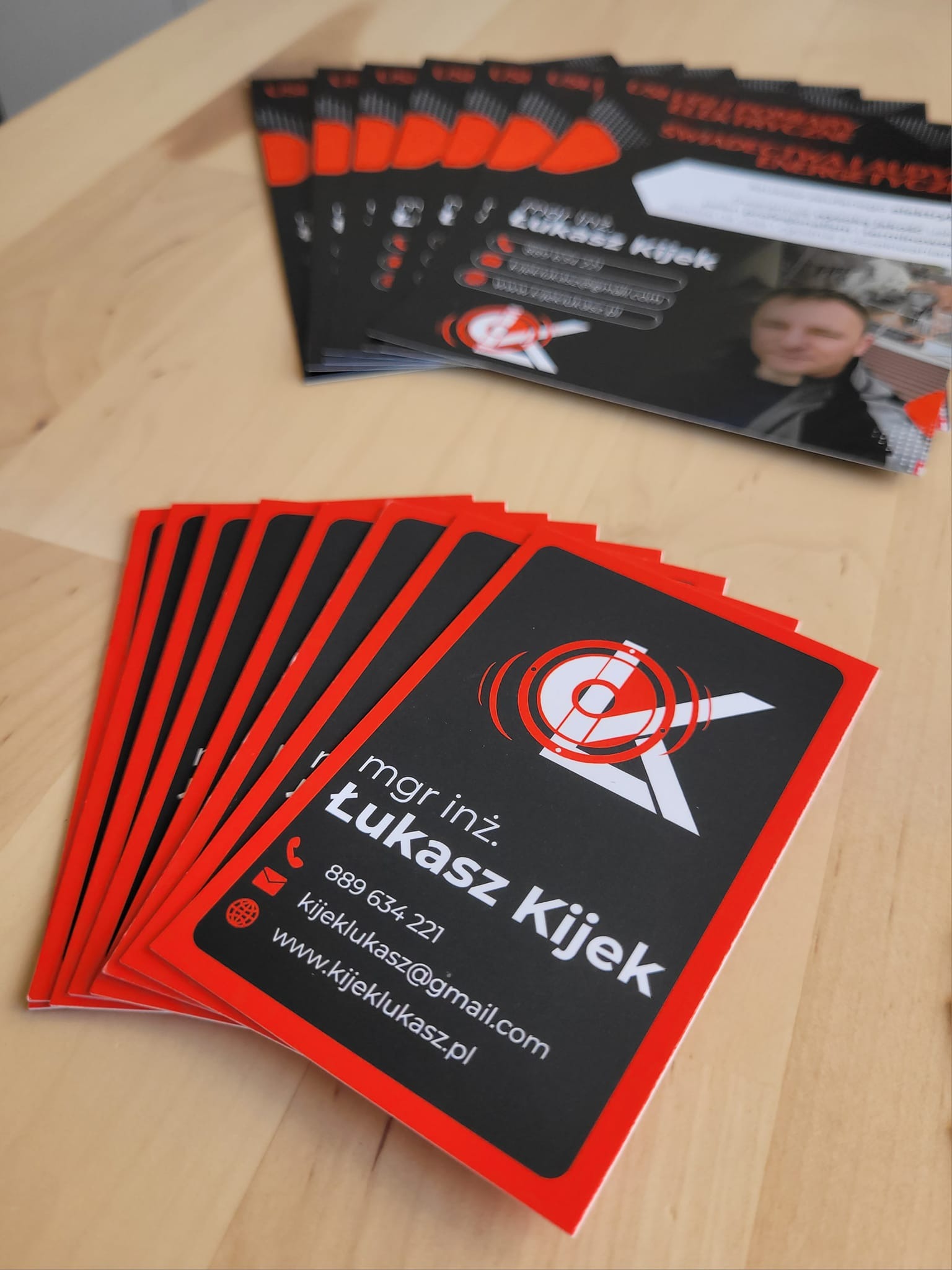 Stos wizytówek mgr inż. Łukasza Kijka z czarnym tłem, czerwonym obramowaniem i białym logo celownika, leżących na drewnianym blacie.