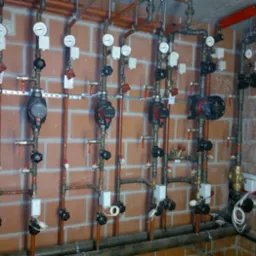 Różnorodne rury, zawory i manometry tworzące instalację grzewczą na tle ceglanej ściany, widoczne pompy obiegowe i elementy sterujące.